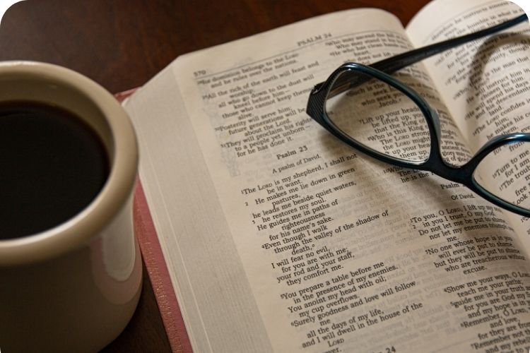 A pair of glasses rest on an open Bible with a mug of coffee to the side.